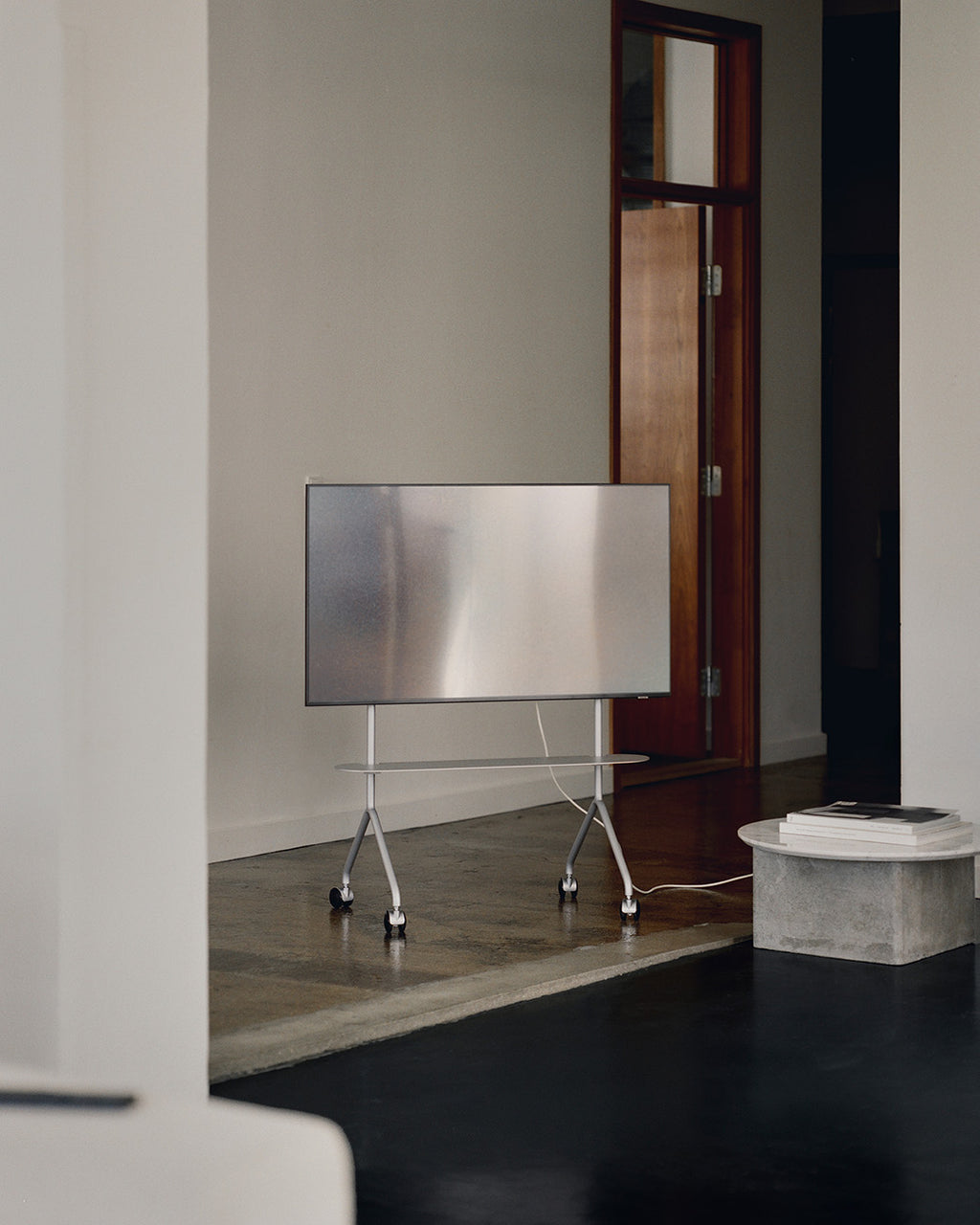 Support TV Moon Light coloris Cloudy Silver sur roulettes, écran monté, dans un intérieur minimaliste près d'une porte en bois